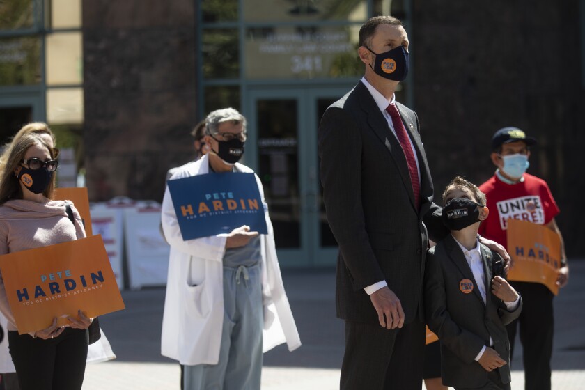 A man stands with half a dozen people. Some hold signs saying "Pete Hardin for O.C. district attorney." All wear  face masks.
