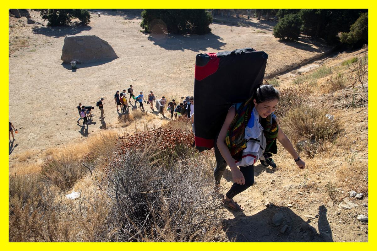 A climber carries a large pack up the side of a hill. A line of folks begins the same ascent below.