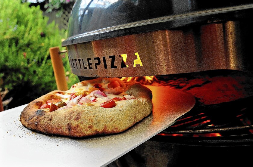 A nicely charred pizza is pulled out of the KettlePizza attachment on the backyard grill.