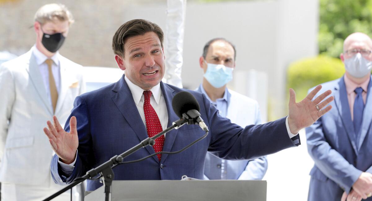 Ron DeSantis speaks into a microphone; several men in masks stand in the background.