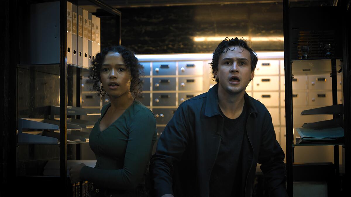 A young woman and man in a vault of safe deposit boxes looking scared