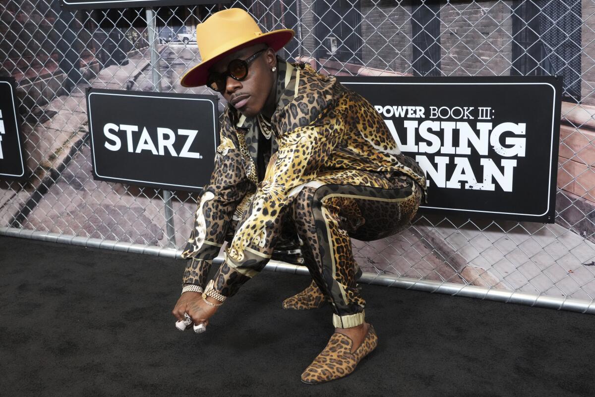 DaBaby, in hat and animal print clothing and shoes, squats as he poses for a photo.