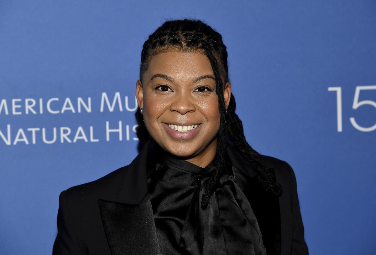 Punkie Johnson smiling in a black shirt, black suit jacket and braids against a blue backdrop