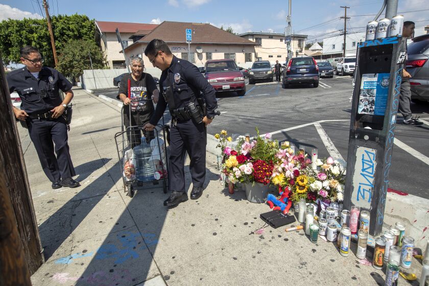 LINCOLN HEIGHTS, CA -SEPTEMBER 12, 2022: LAPD officers Nicholas Torres, left, and Erick Rodriguez help a blind man (didn't want to give his name) who lives in the neighborhood, make his way past a memorial for Winfield Mikey Lee, 17, on Workman St. in Lincoln Heights, one two teenage boys who were shot and killed Sunday night at a street carnival in Lincoln Heights.The LAPD officers were keeping an eye on activity around the memorial. (Mel Melcon / Los Angeles Times)