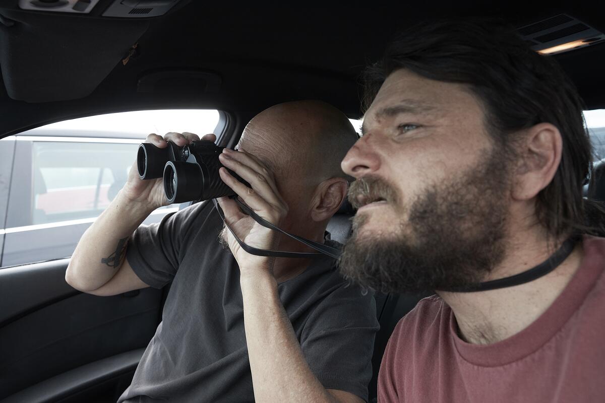 Two men in the front seat of a car, one looking through binoculars.