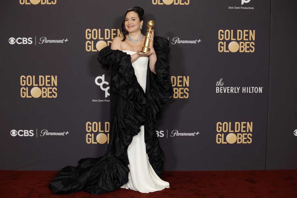 Lily Gladstone wears a white dress and long black shawl and holds up her Golden Globes award as she poses for photos