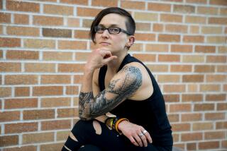 A woman with straight hair in tank top with many tattoos in front of a brick wall.