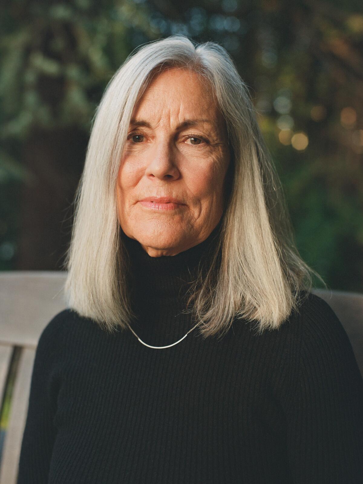 A woman with long, gray hair in a black turtleneck sitting on a white bench