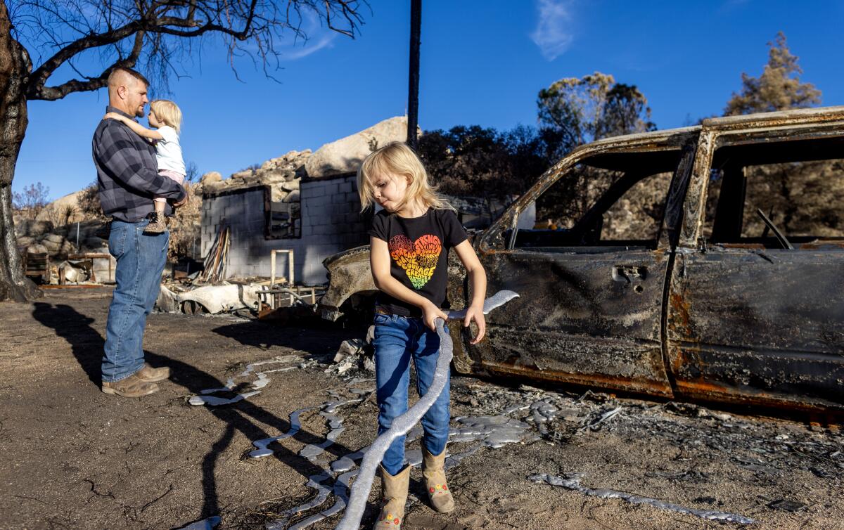 A man holding a small child stands with a girl next to the burned hulk of a car.