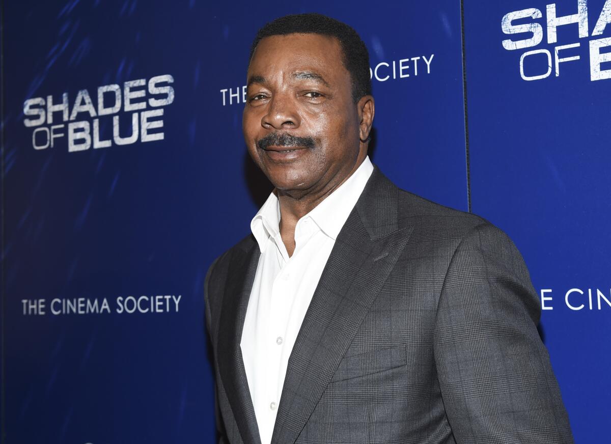 Carl Weathers in a gray blazer and white shirt standing in front of a blue backdrop with the words "Shades of Blue" on it.
