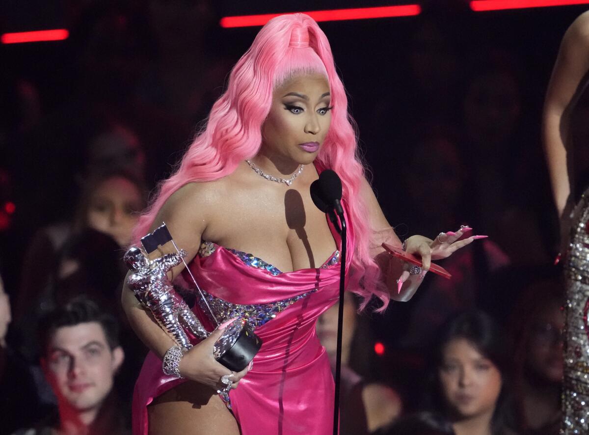 Nicki Minaj in a hot pink gown speaks into a microphone and holds a silver trophy in the shape of a man in a moonsuit