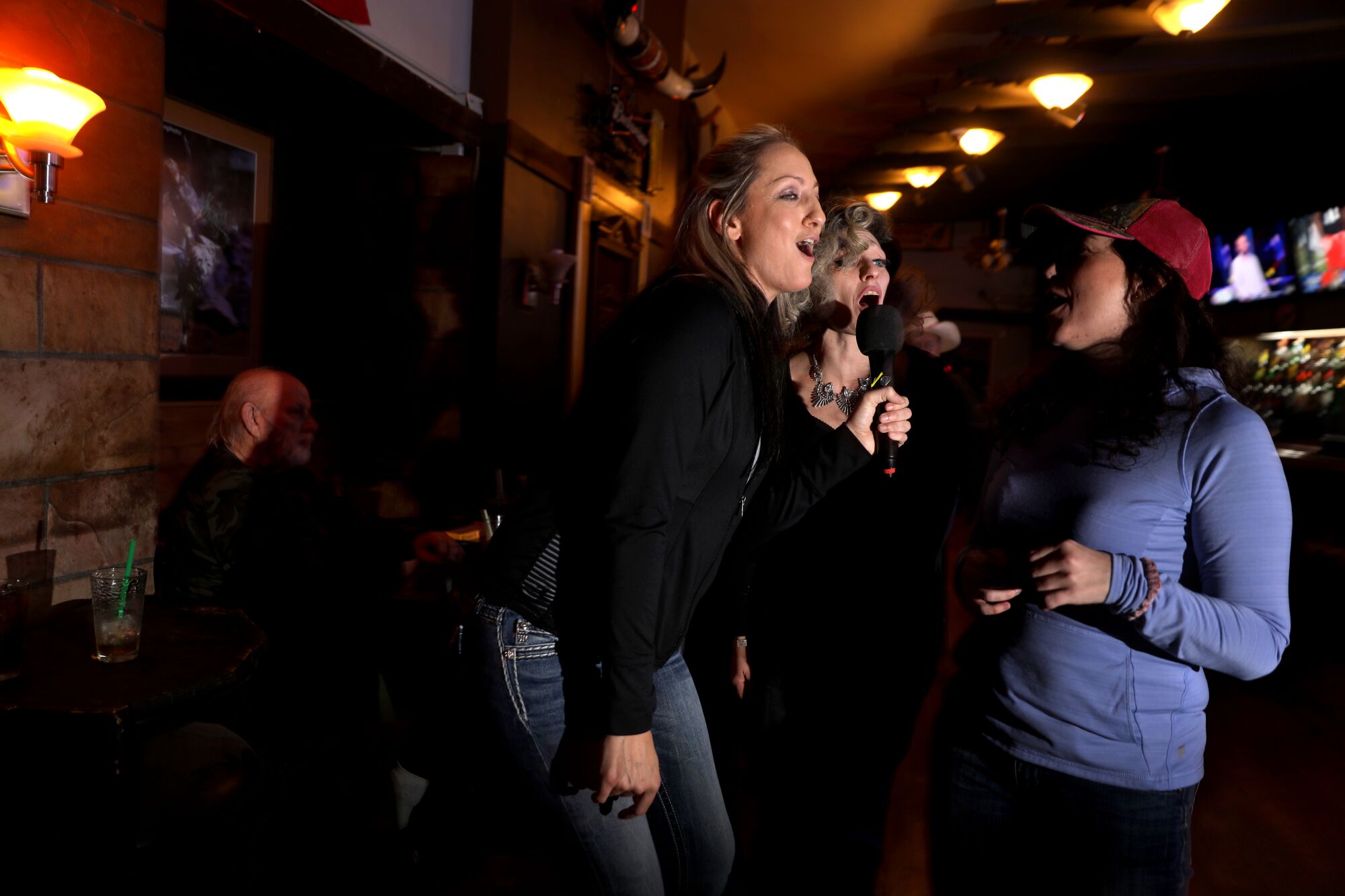 A group of women, none with masks, lean in to sing karaoke around one microphone in a bar
