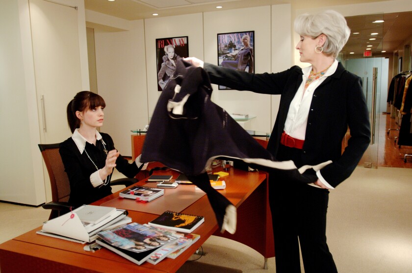A woman with white hair hands a garment across a desk to her seated young female assistant.