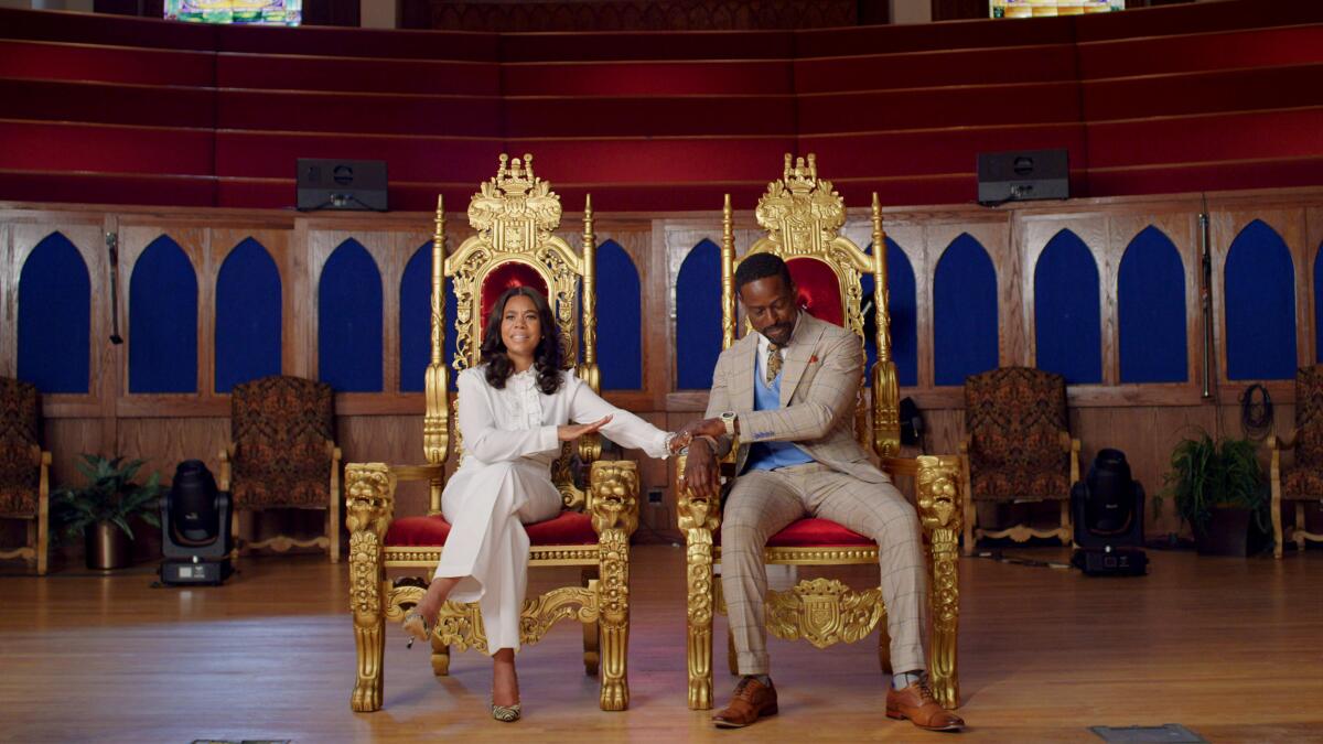 A woman and man sit on matching golden thrones upholstered in red velvet.