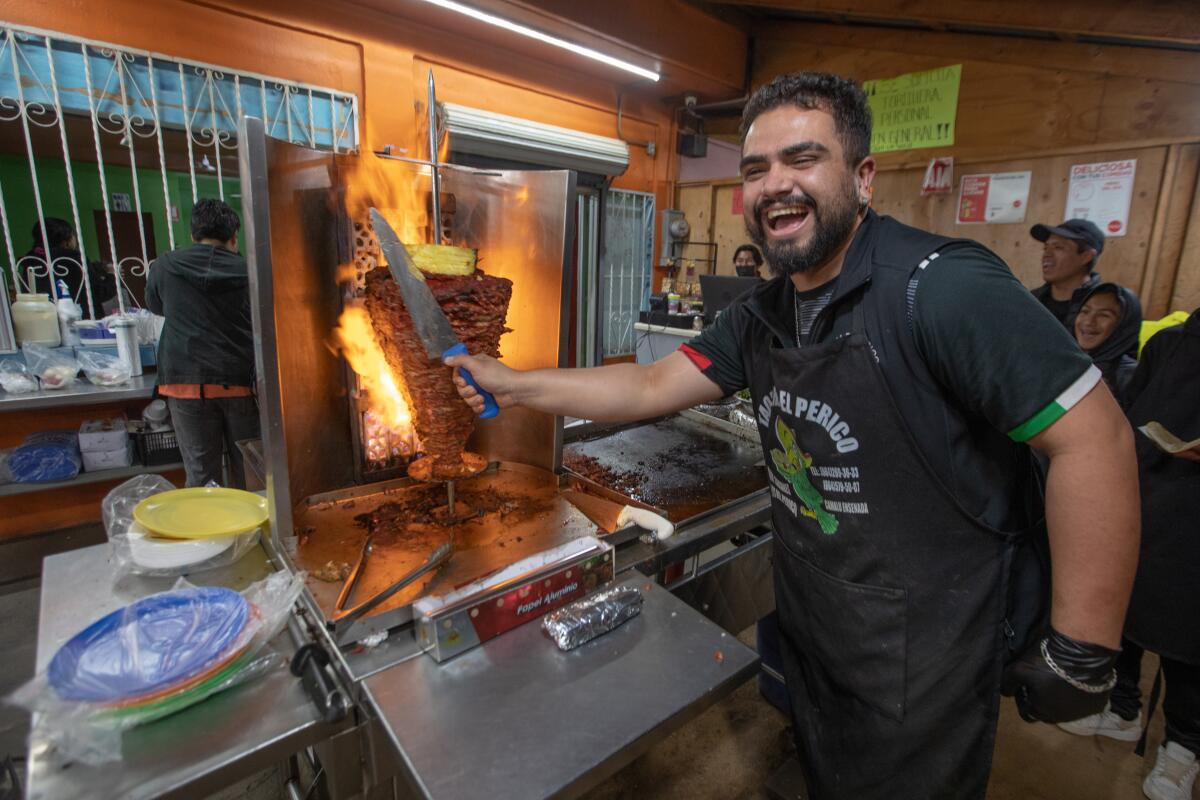A man stands with a knife in a kitchen, cutting meat off a flaming spit and grinning.