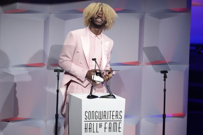 A man with curly blond hair wearing a tuxedo stands on stage