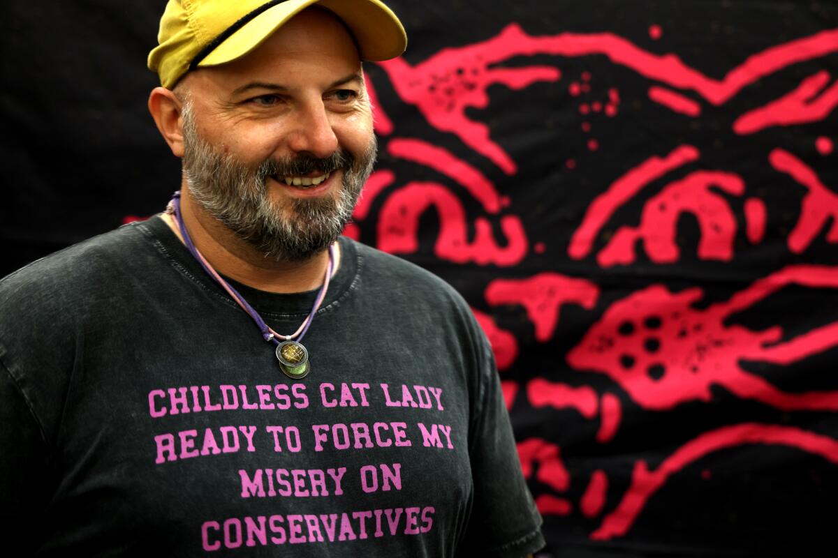 A man smiling and wearing a T-shirt, a baseball cap and necklaces in front of an illustration of a cat.