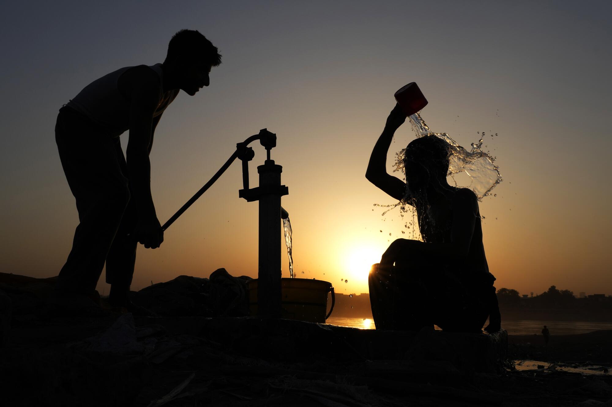 The setting sun silhouettes two figures at a water pump as one of them ours a container of water over their head.