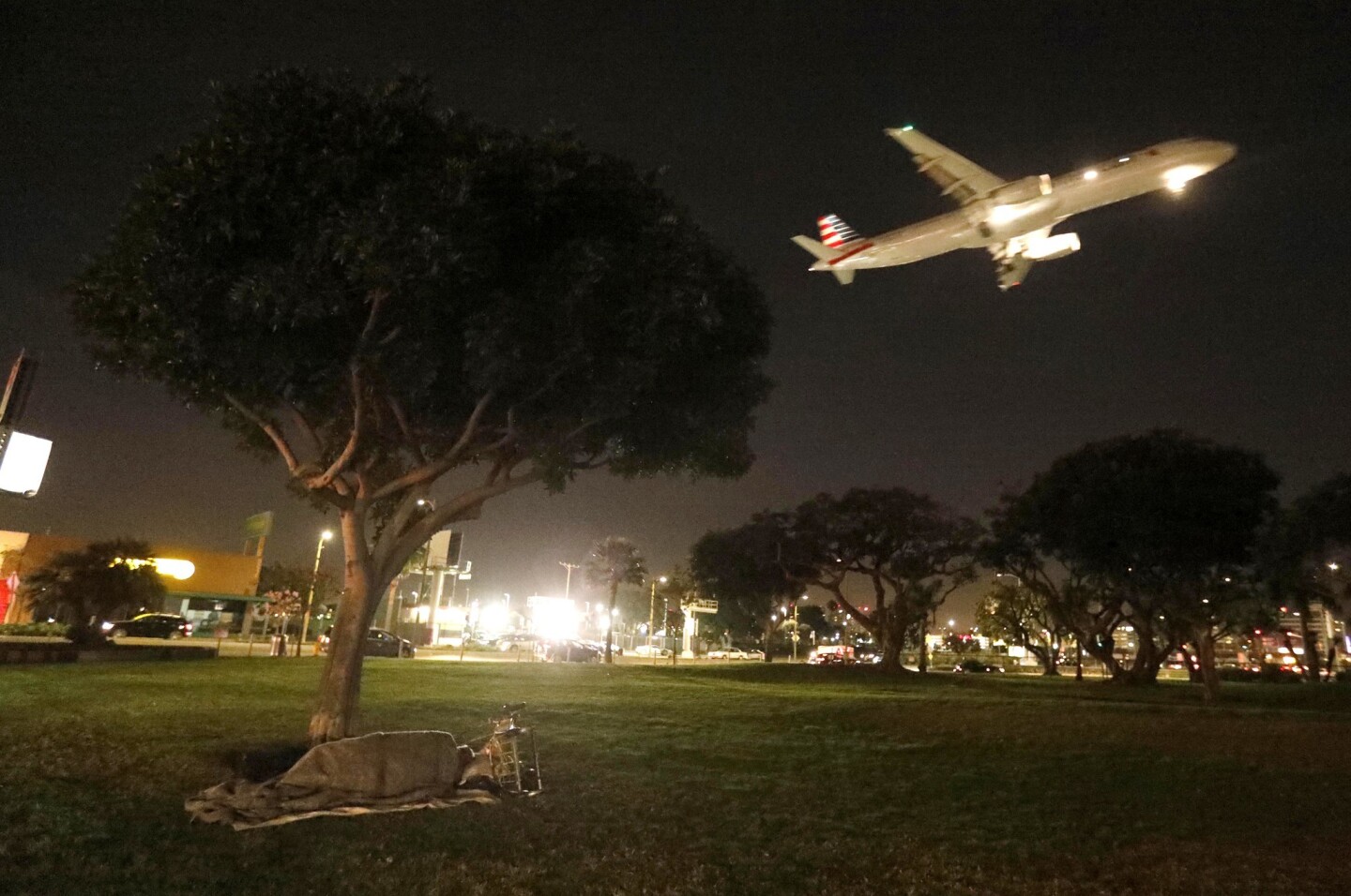 A homeless person sleeps in a park at the corner of W. 92nd Street and Sepulveda Blvd. as a plane comes in for a landing at the Los Angeles International Airport in Westchester.
