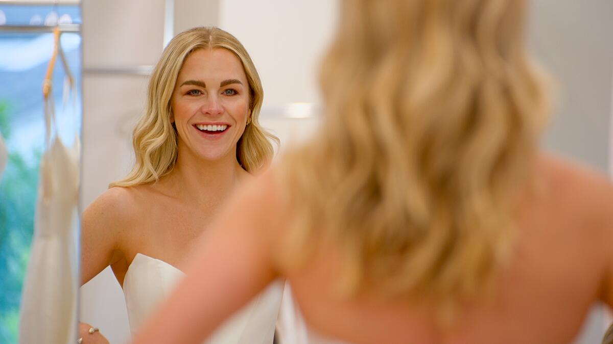 Seen from the waist up, a blond woman in a strapless white wedding dress smiles at herself in the mirror.