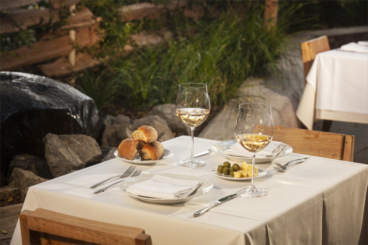A photo of a table for two with poured glasses of white wine and a plate of olives with butter, and a basket of bread.