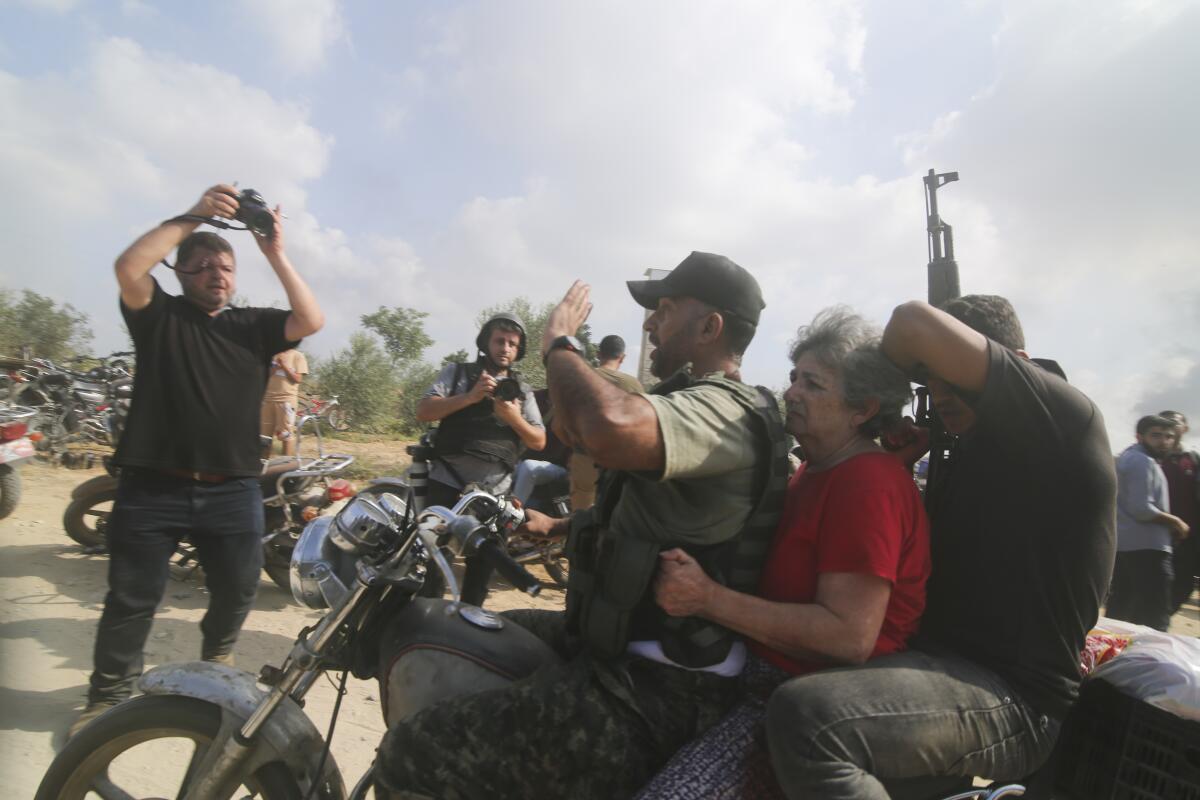 A woman sitting on a motorcycle between two men, one holding an assault-style rifle, as a man takes photos and others look on