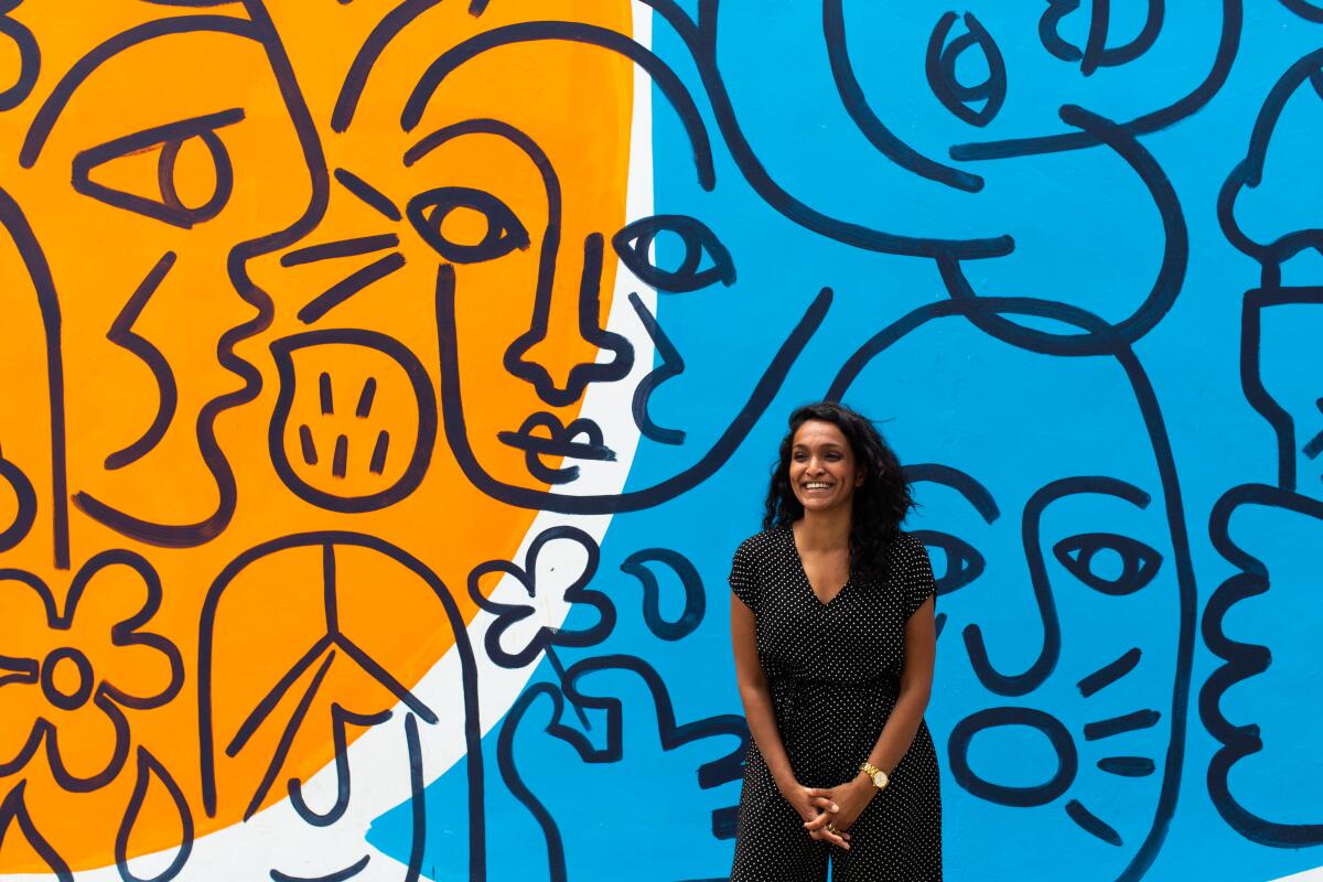 A woman in a short-sleeved jumpsuit stands in front of a colorful wall mural.