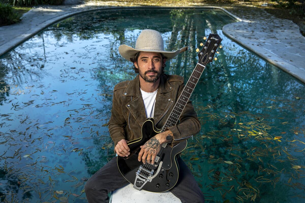 A man holding an electric guitar sits in front of a swimming pool