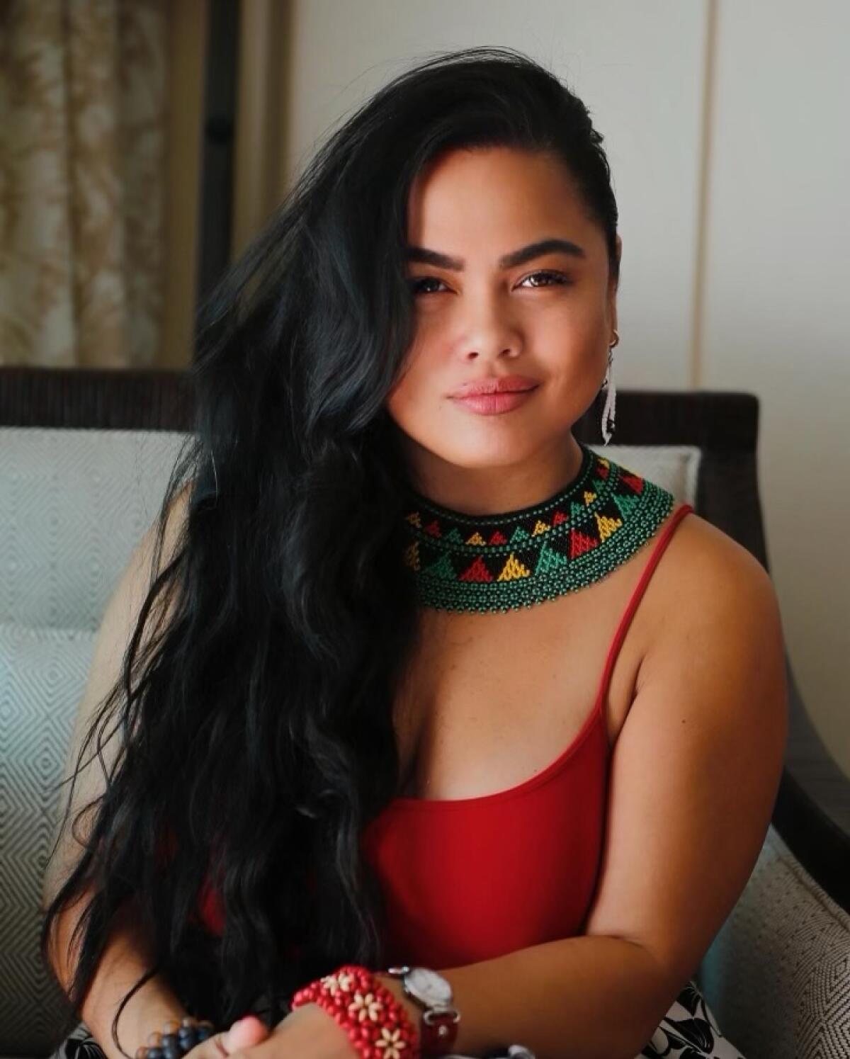Roslynn Alba Cobarrubias smiles with her hands folded while wearing a red tank top and a colorful patterned necklace