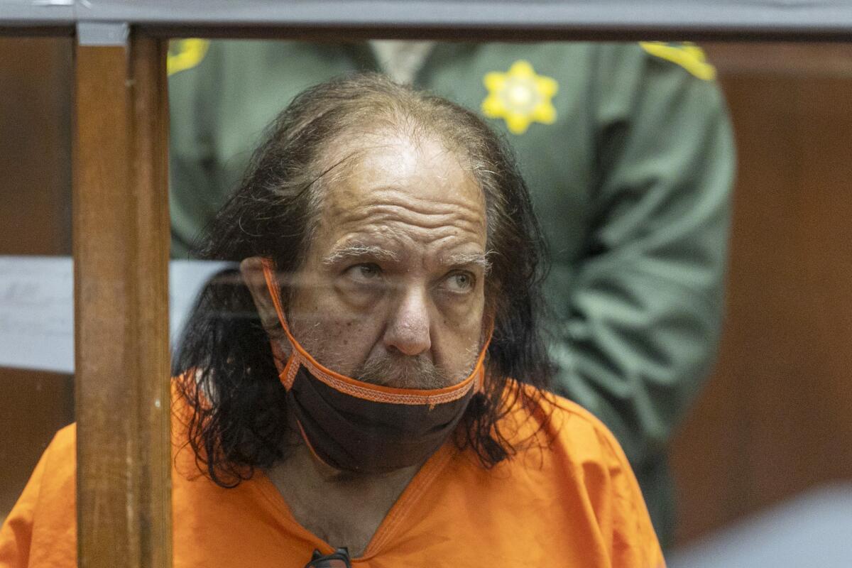 Ron Jeremy sits in a courtroom wearing an orange jail jumpsuit and a mask