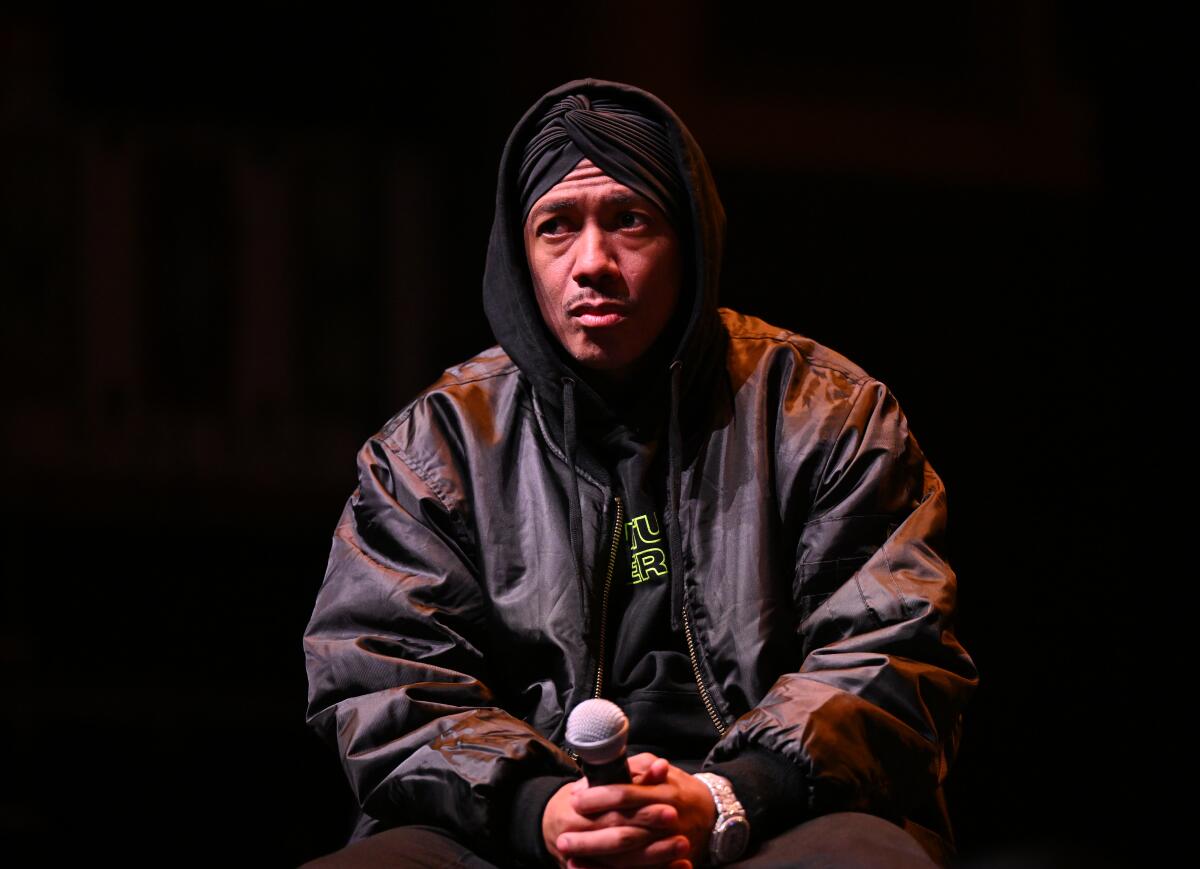 Nick Cannon wearing a black hoodie and jacket and holding a microphone on stage
