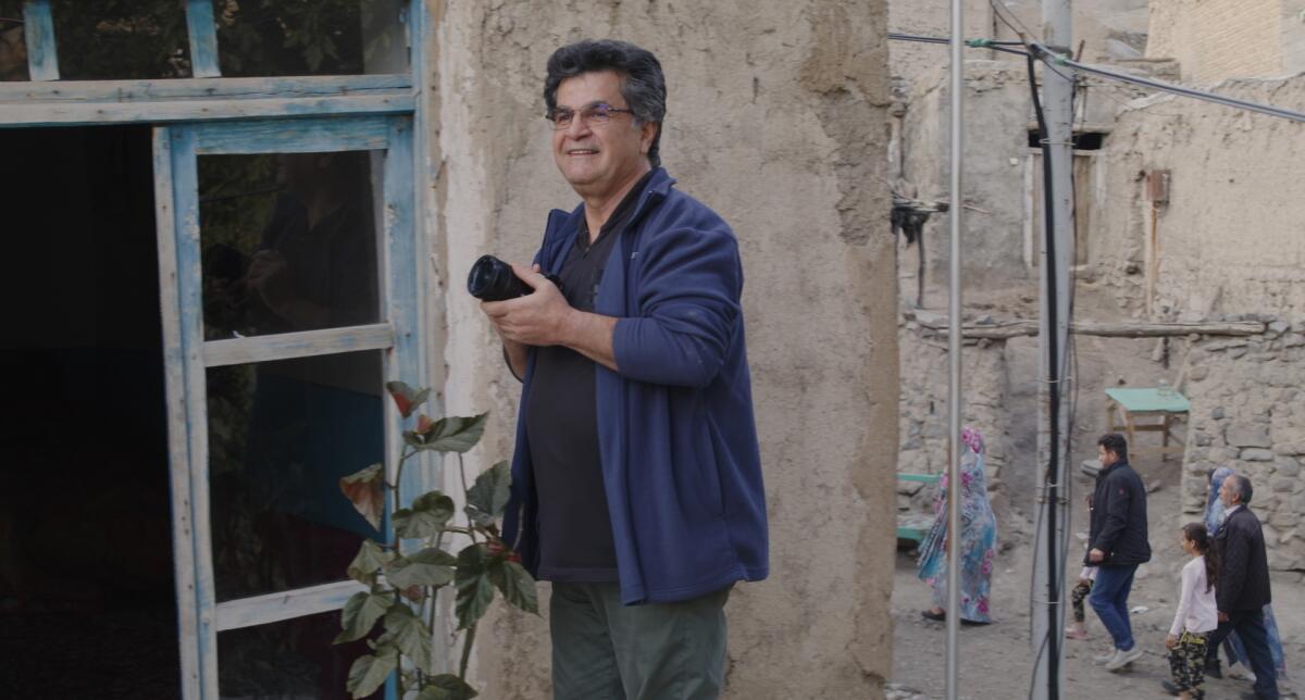 Jafar Panahi holds a camera while people walk in the background.