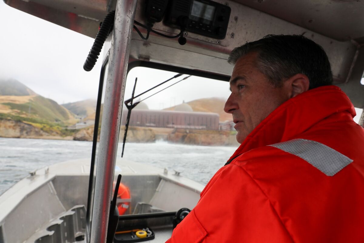 A man in orange jacket pilots a boat; the shore can be seen in the distance.