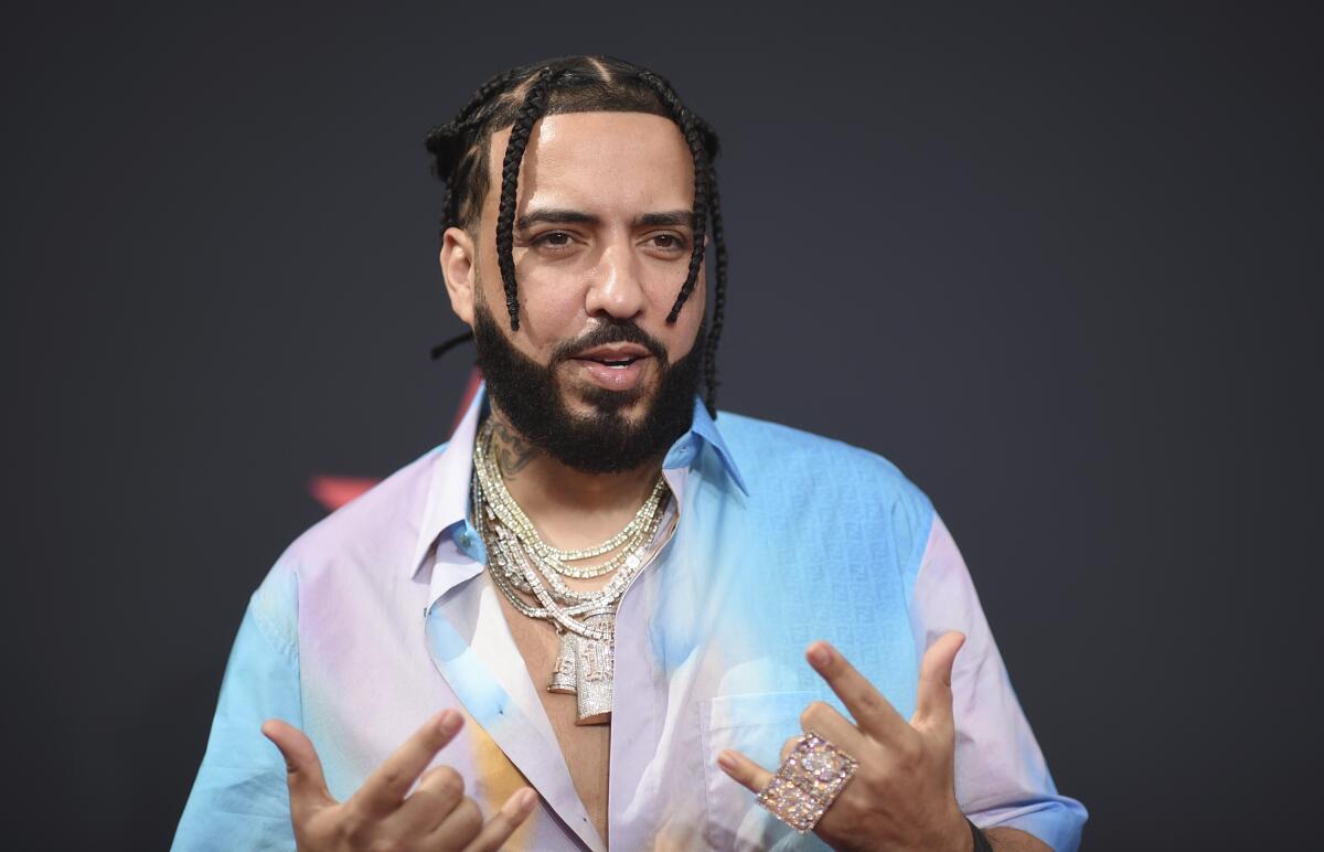 A man with braids, a beard and gold chains over a multicolored shirt makes gestures with both hands.