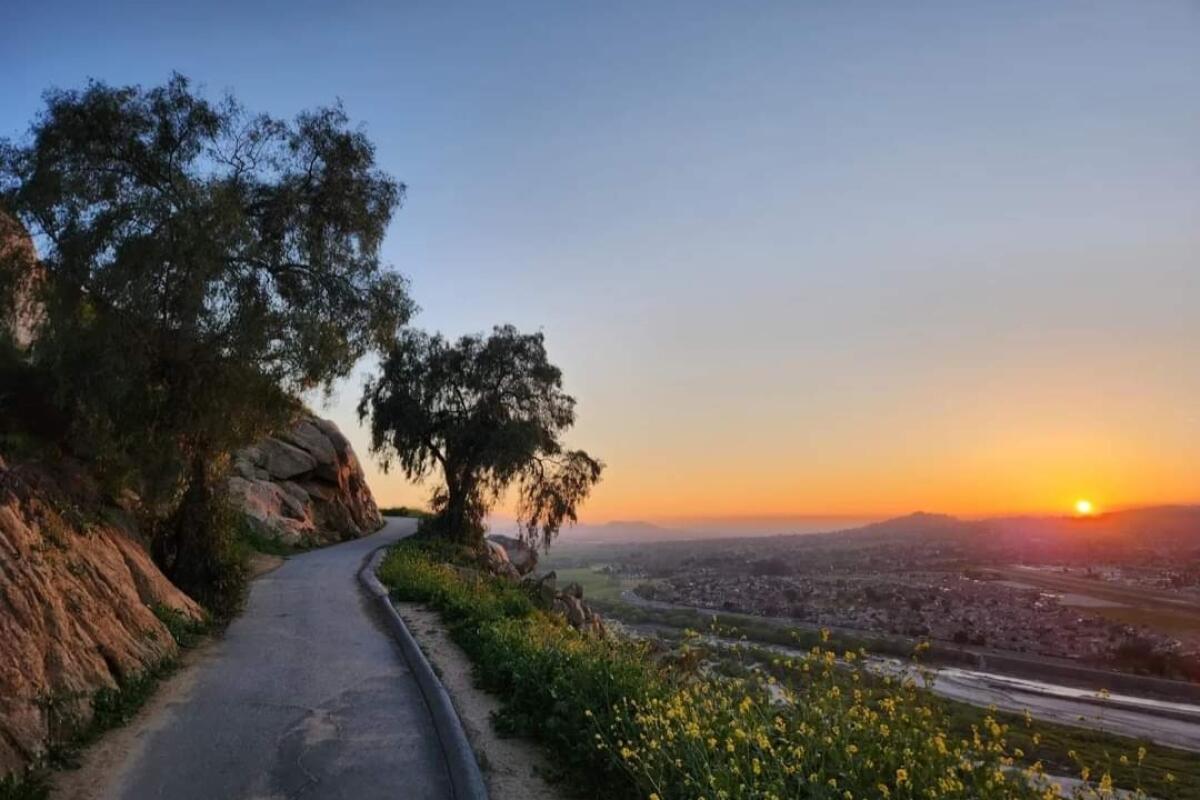A hiking trail winds along a mountain side with a sunset and wide view of a city to the right.