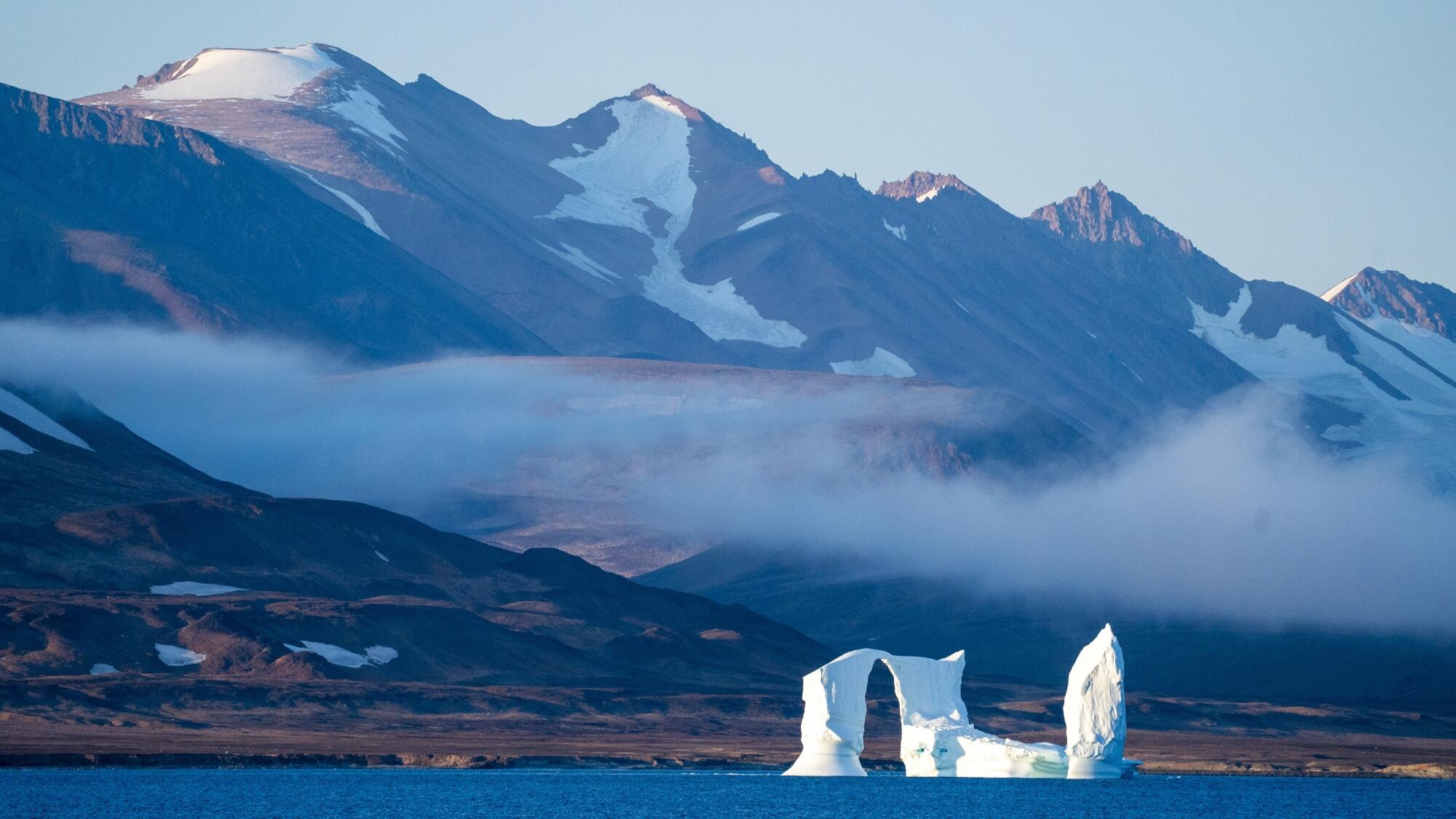 An iceberg floats in front of snowy mountains.