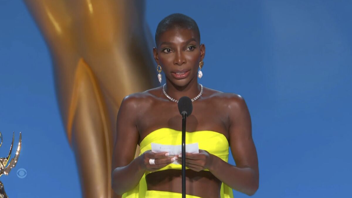 A woman in a bright yellow dress holds a piece of paper and speaks into a microphone
