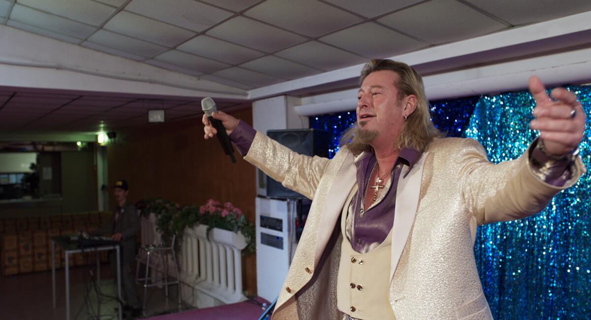 A man in a white three-piece suit with a shaggy goatee and hair stands with his arms spread wide, a microphone in one hand