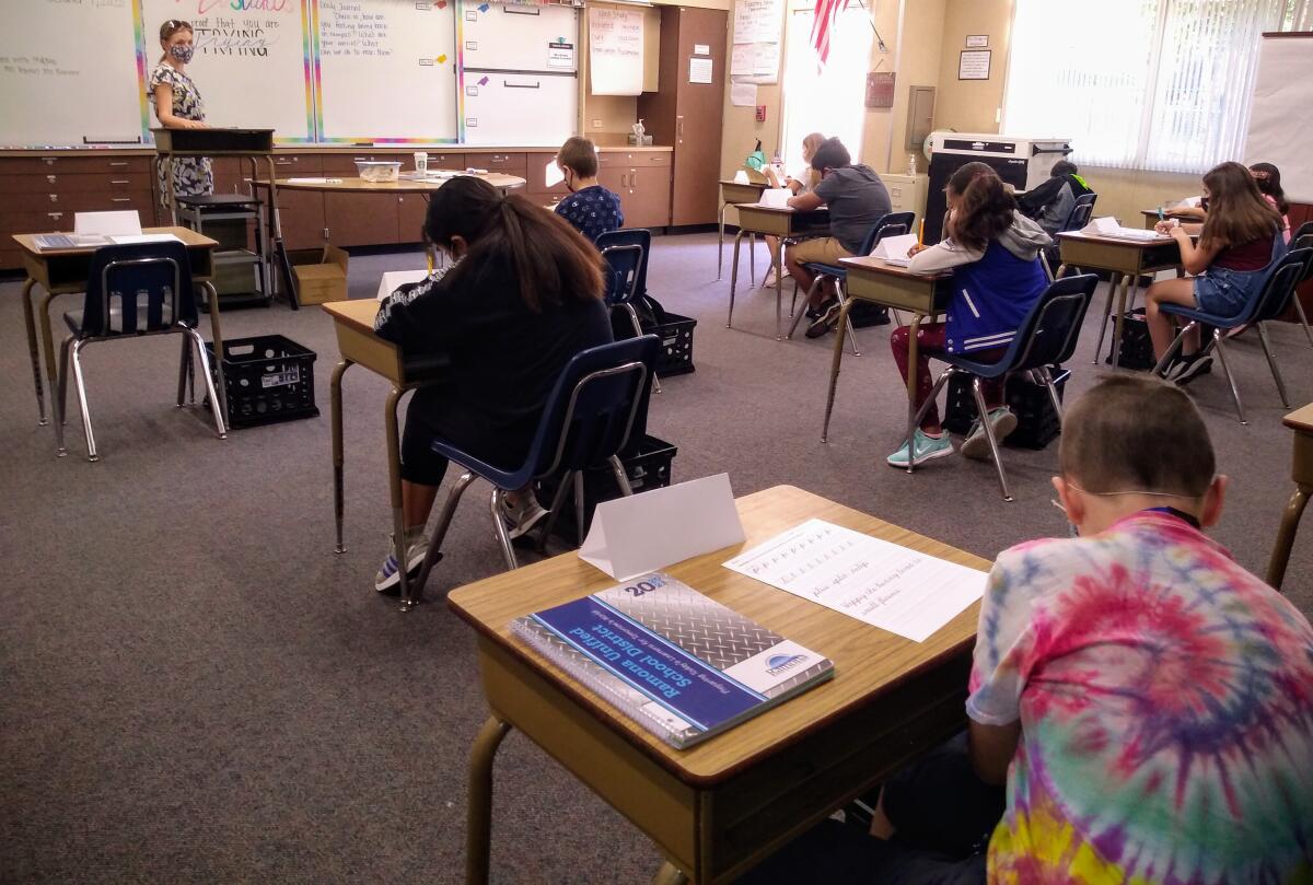 Inside a classroom, children sit at desks spaced apart. A teacher in a mask stands at the front of the class.