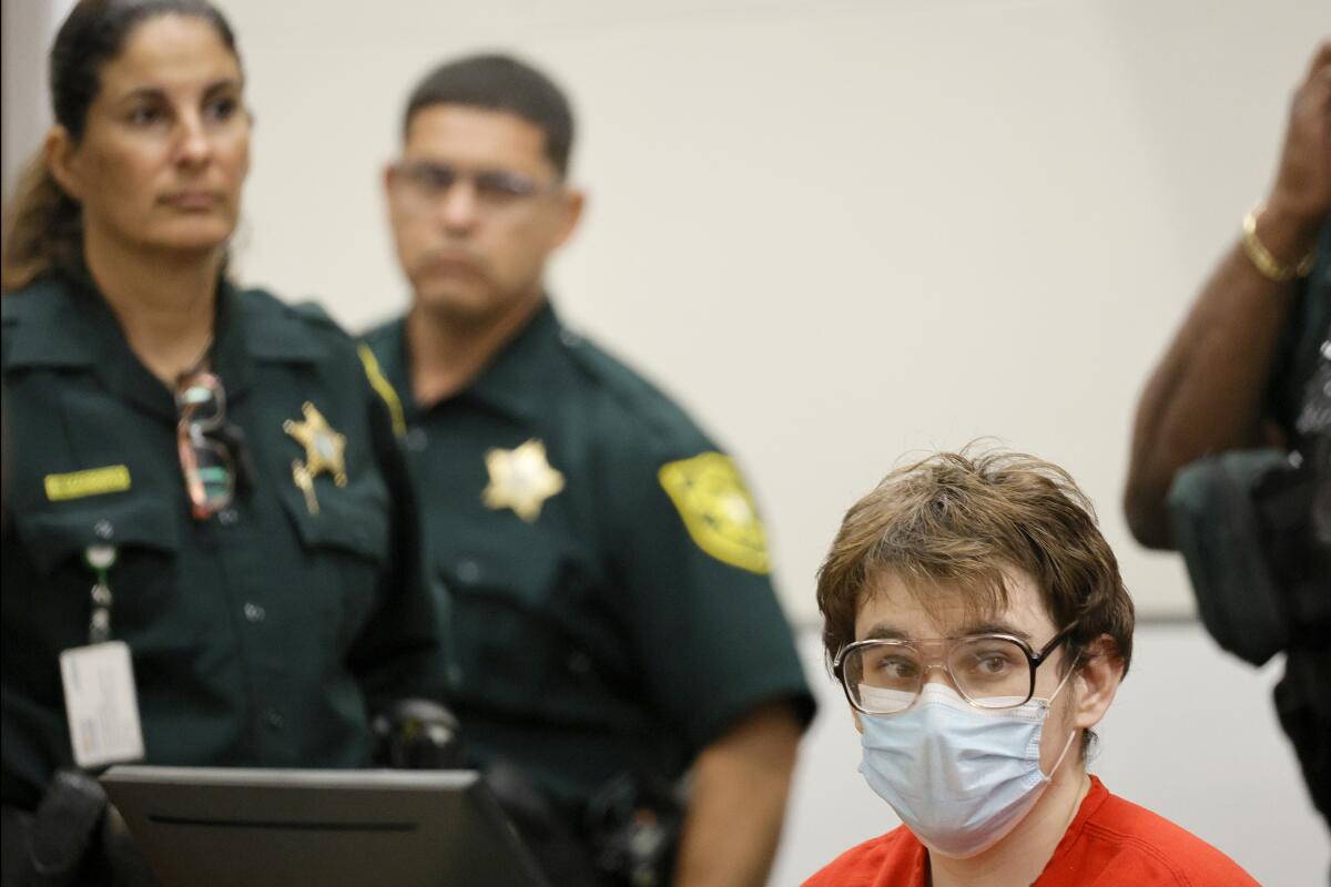 Nikolas Cruz sits in court as law enforcement officers stand nearby.