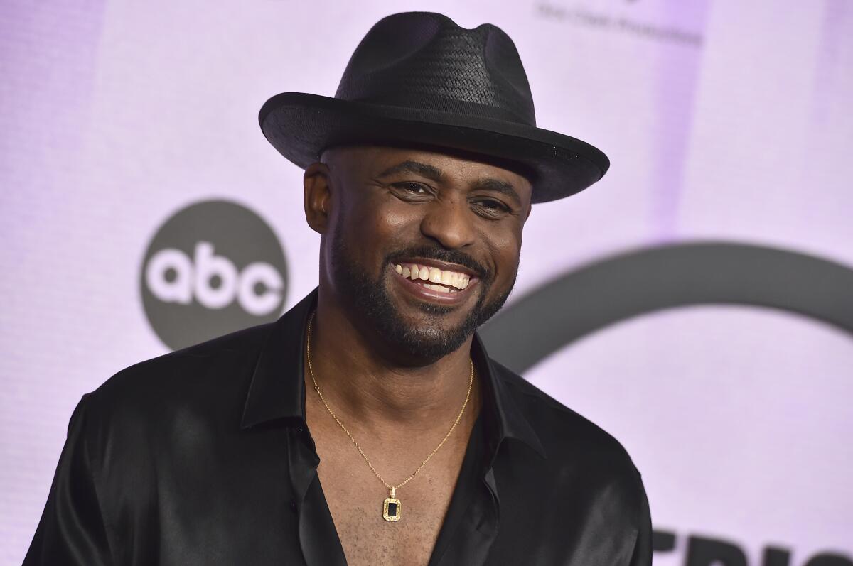 Wayne Brady smiles while clad in a black fedora and a black button-up shirt revealing his chest and a gold necklace