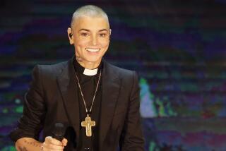 Sinead O'Conner smiling while on stage, holding a mic in one hand, wearing a black suit, priests collar and golden cross