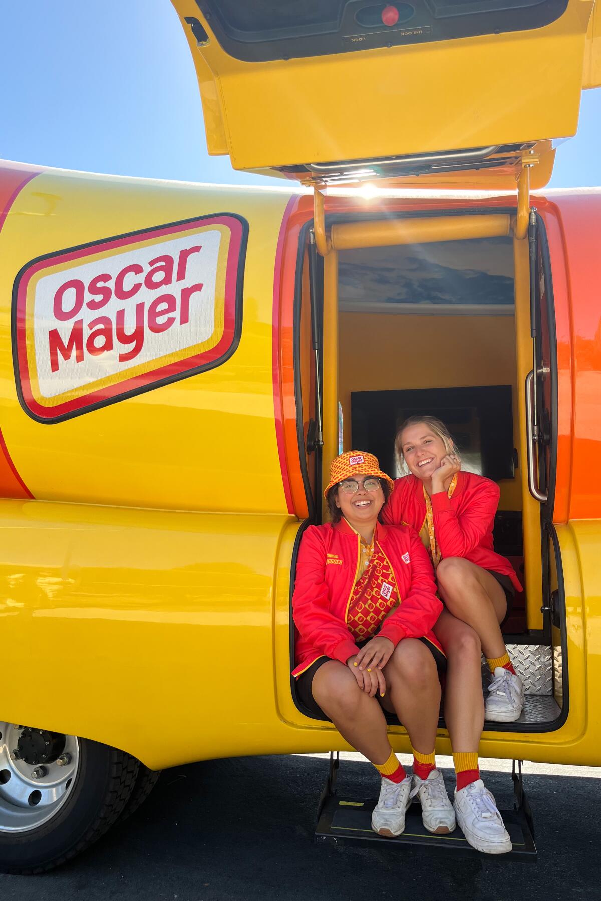 Two women sit on the steps of an open side of the Oscar Mayer Wienermobile.