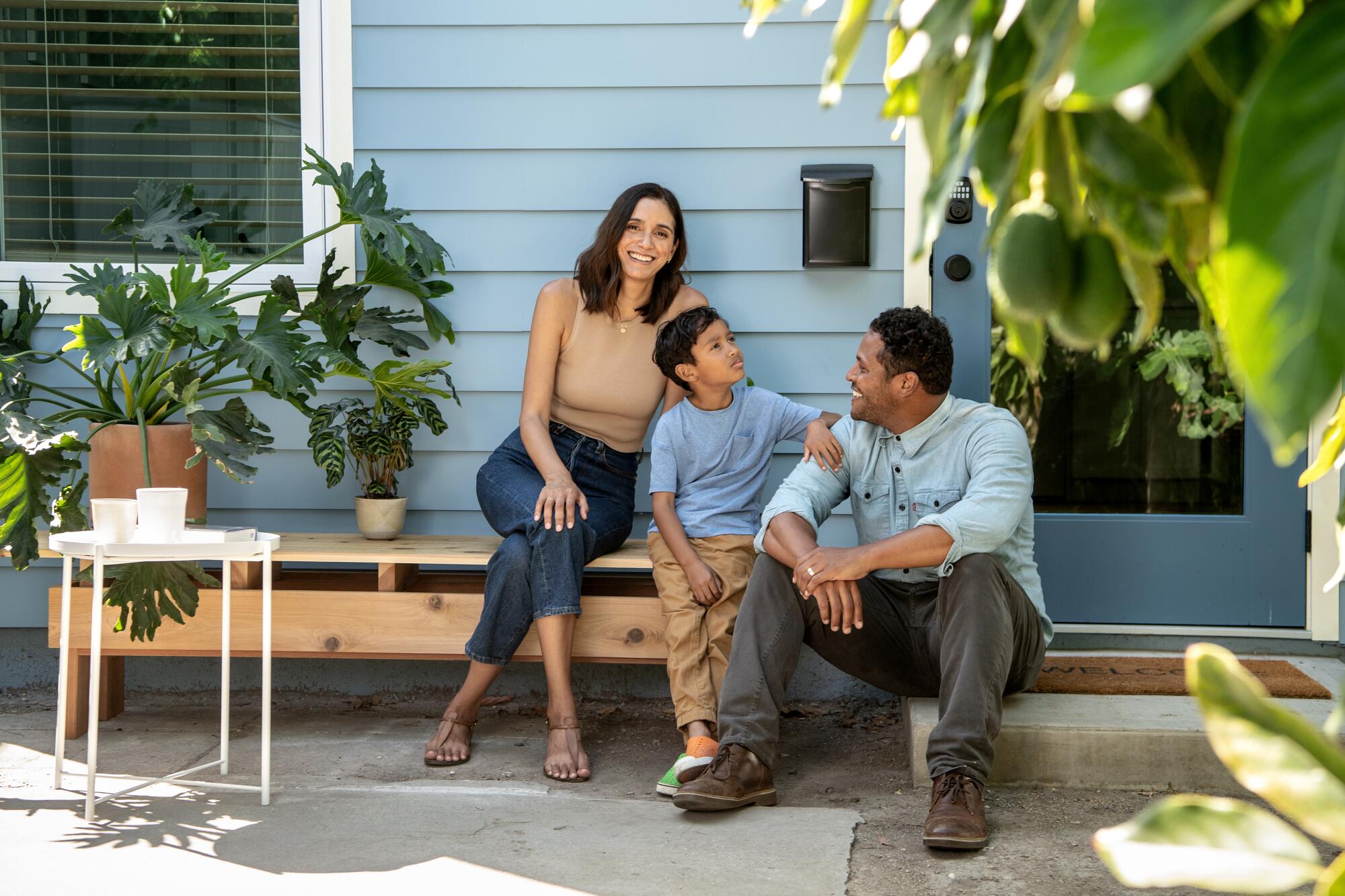 A woman, boy and man sit amid potted plants outside a house with blue clapboard siding 