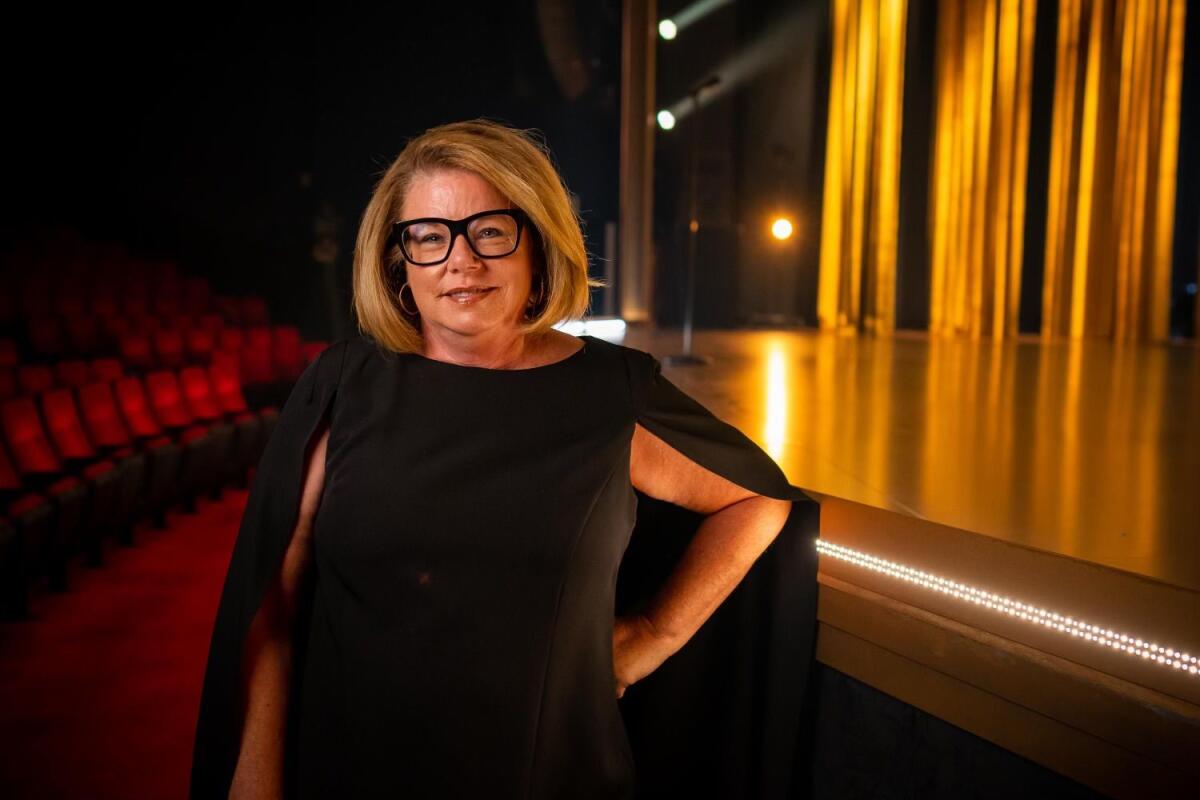 A woman in a black cape dress wearing black-framed glasses and a blond bob stands in an empty theater