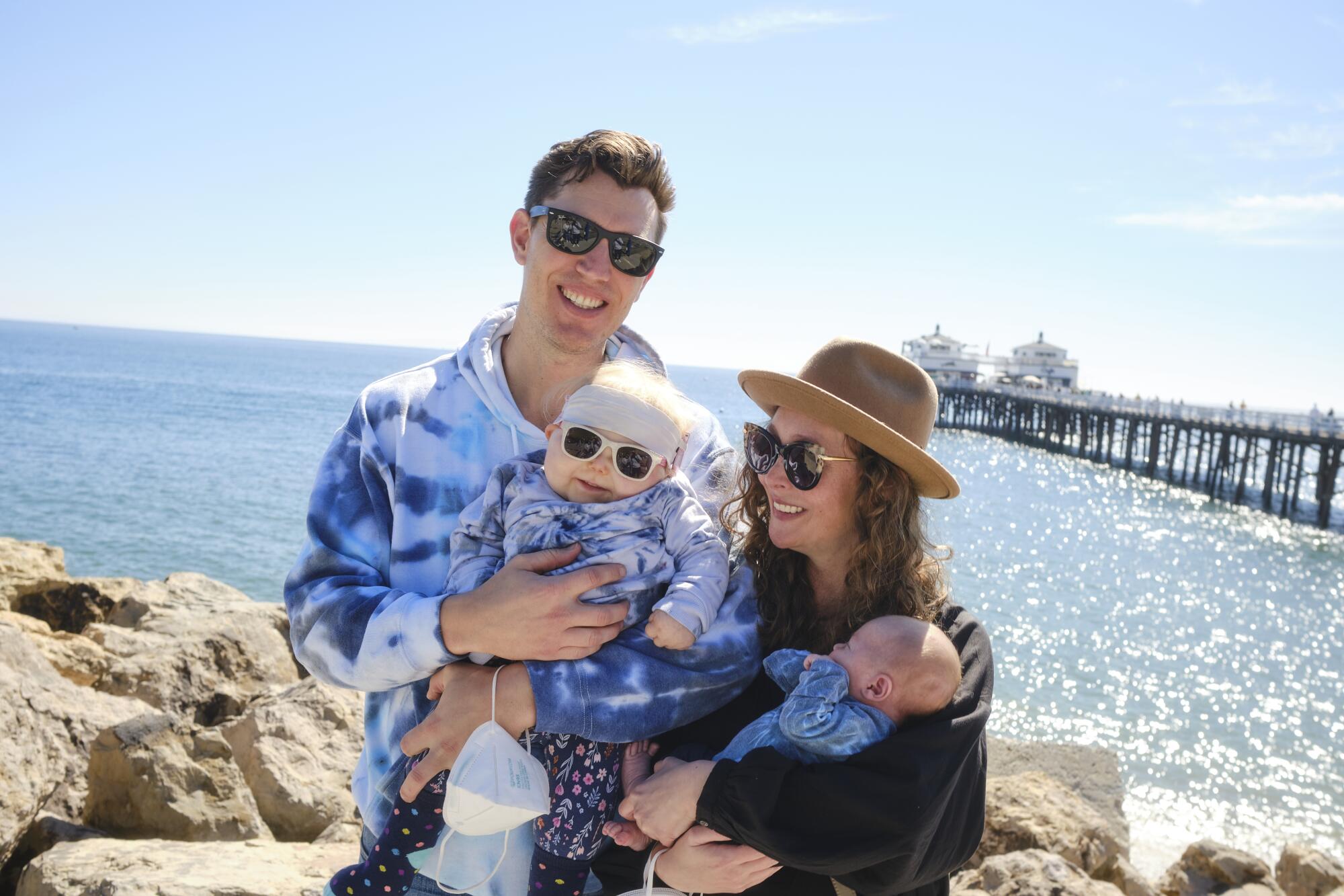 A man and a woman, each holding a small child, on a beach with a pier and the ocean behind them.