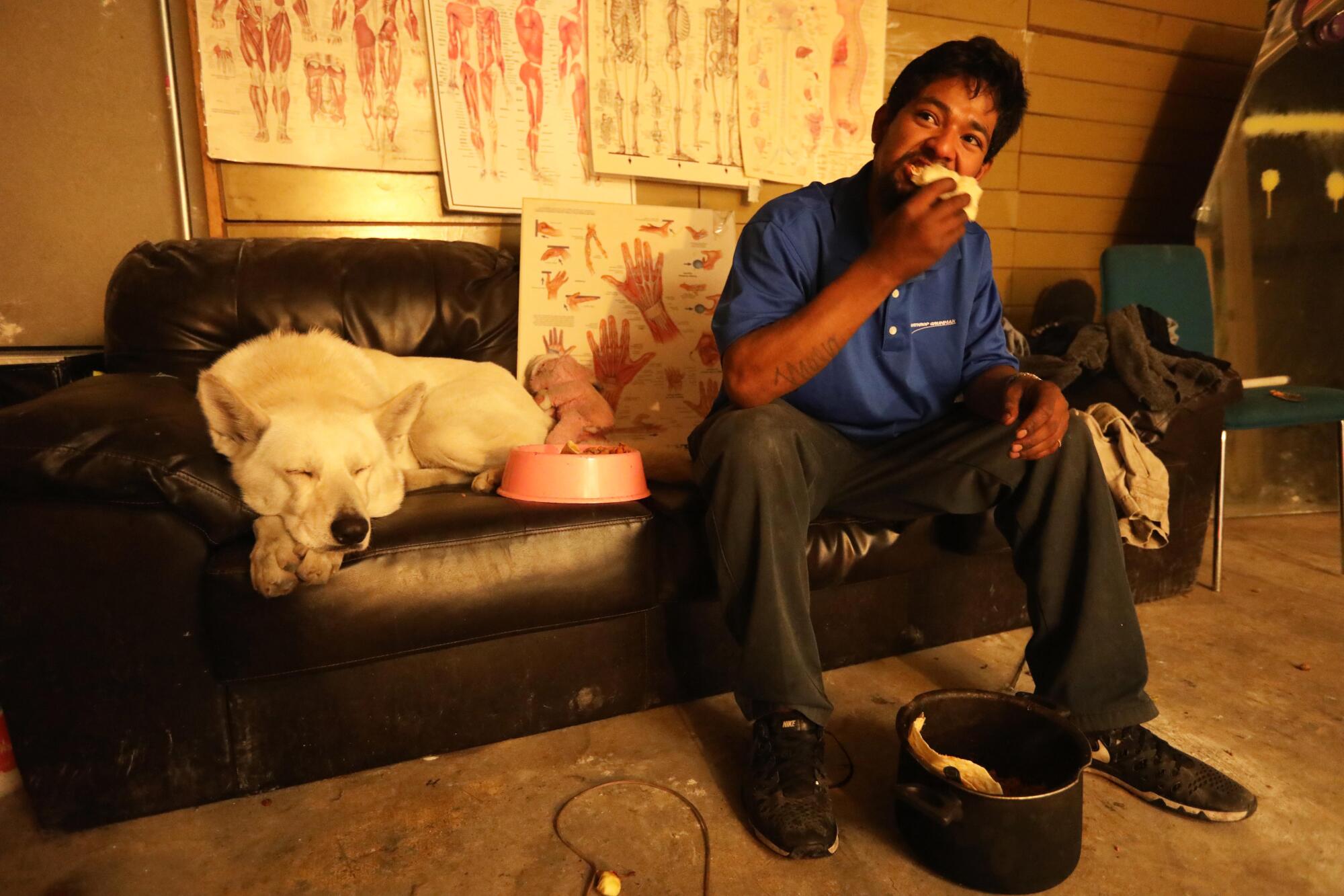 A man eats while sitting on a couch next to his dog and a dog food bowl