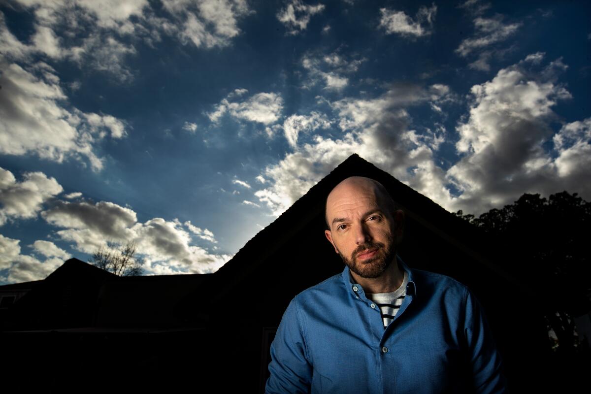 Paul Scheer stands in front of his house, its outline in black in the background, under a sky full of clouds.