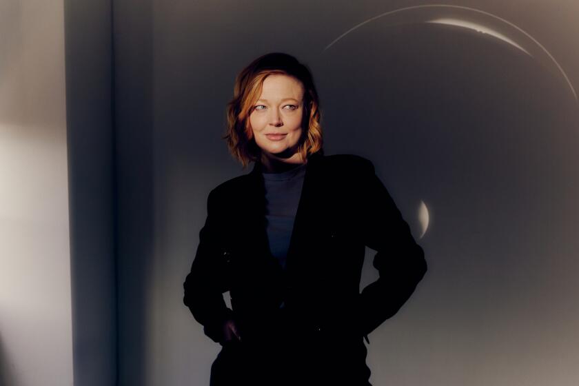 Sarah Snook stands in front a blank wall with her hands in her black blazer pockets and her eyes looking to the side