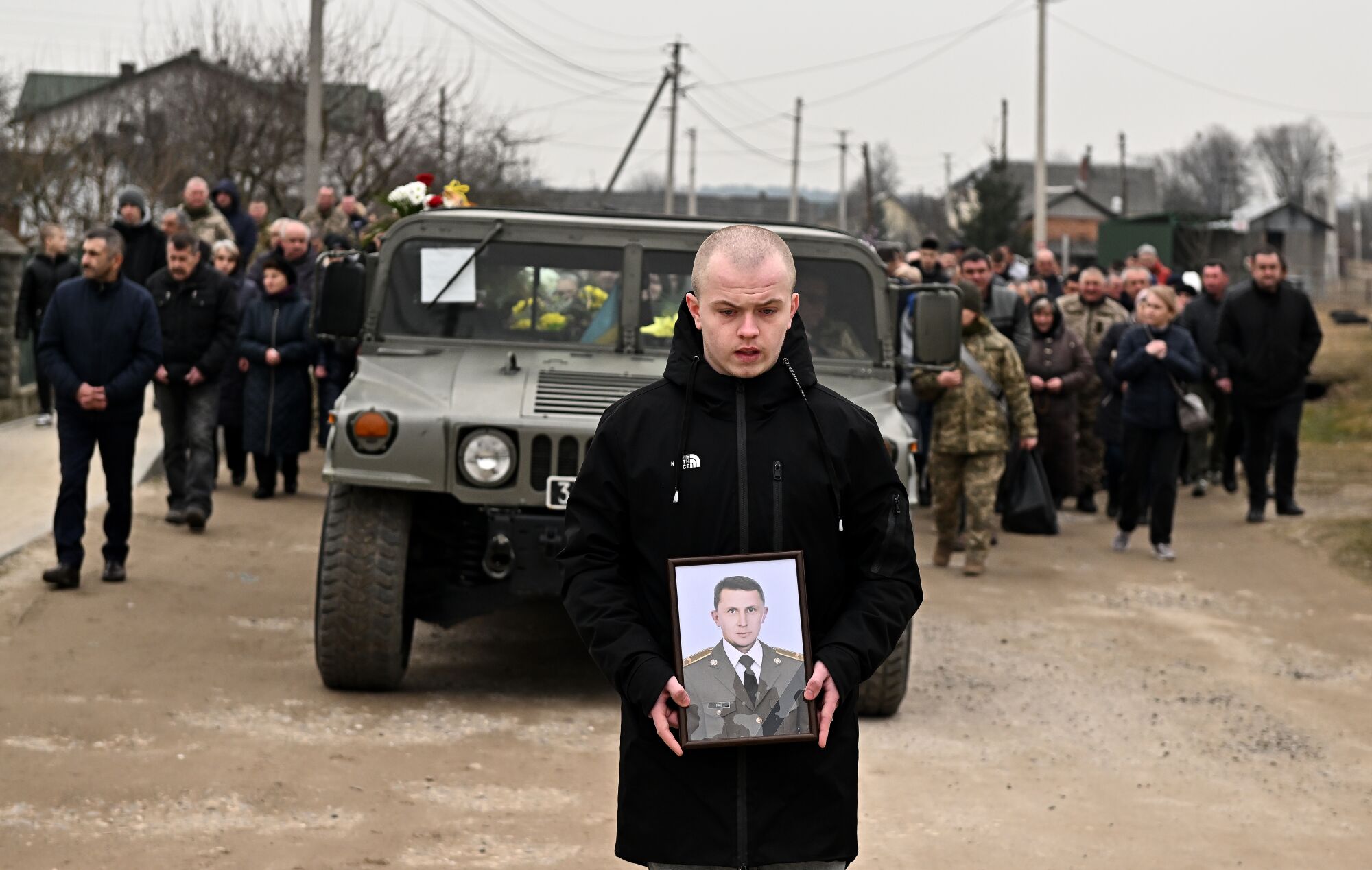 A man carries a portrait in front of a vehicle and a procession of people.